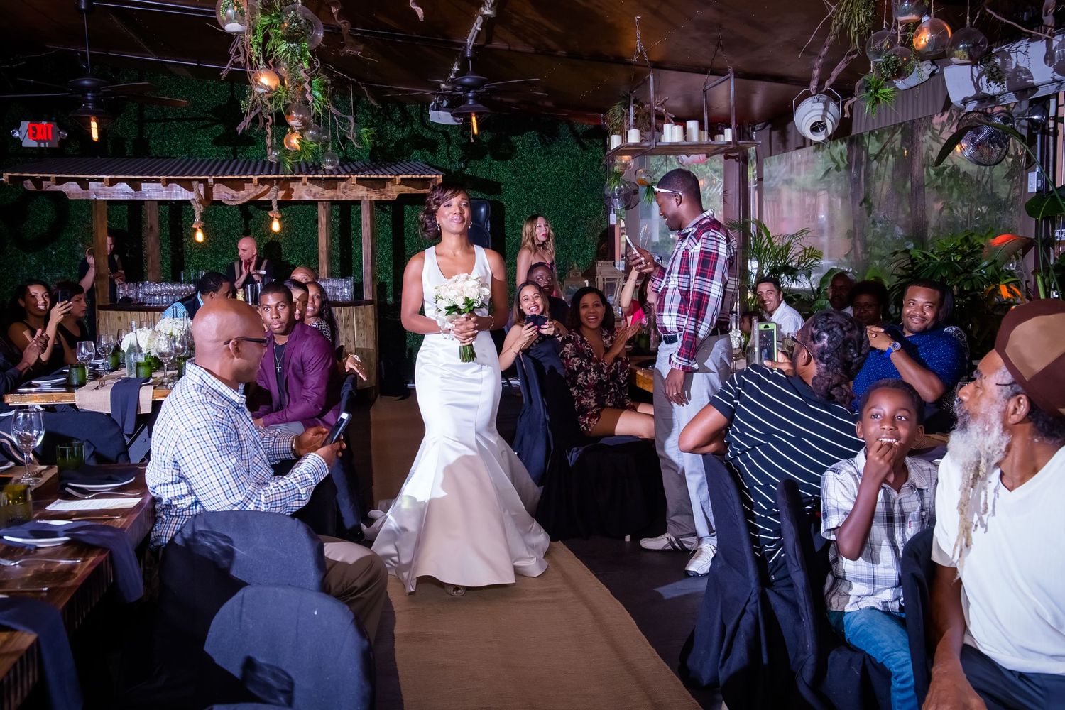 Bride in mermaid dress walks down aisle while guests watch in an intimate venue setting.
