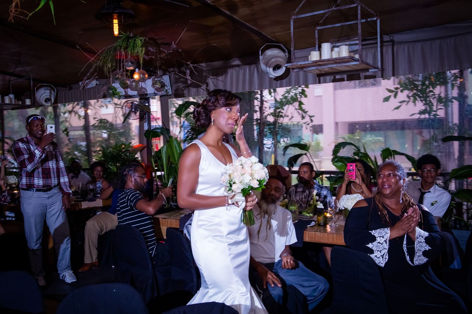 A bride in a fitted white gown walks through an intimate indoor wedding venue.