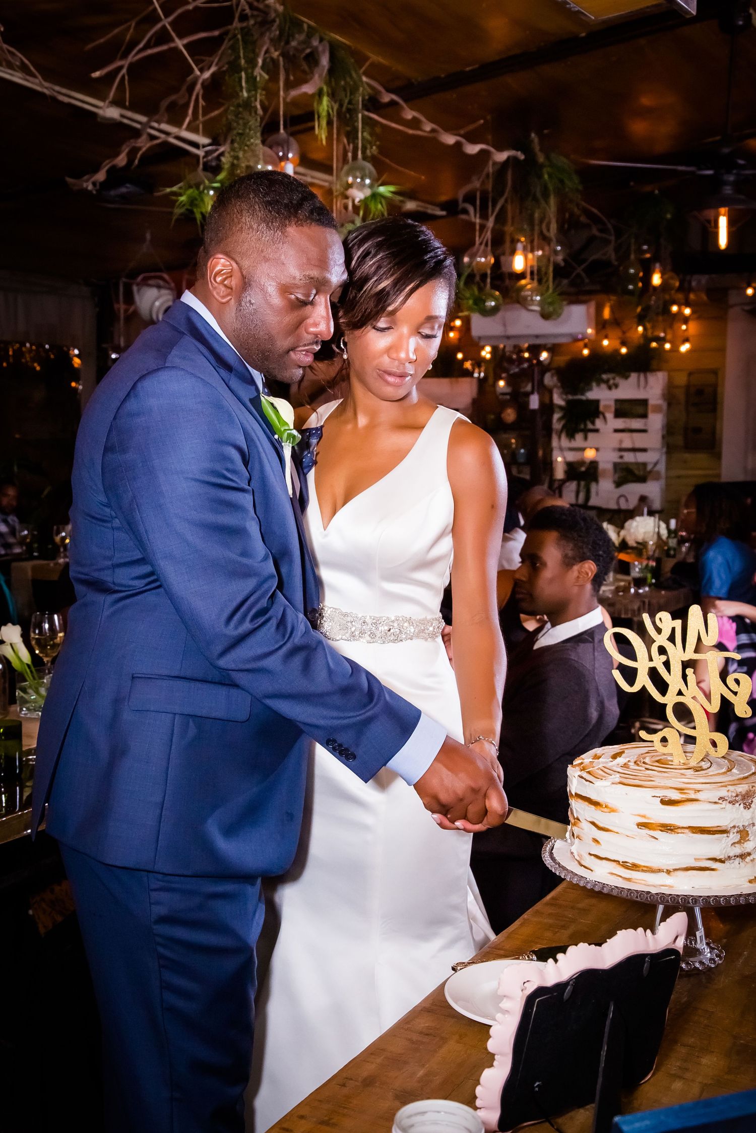 Wedding couple sharing a sweet moment while cutting their tiered white cake at an indoor reception.