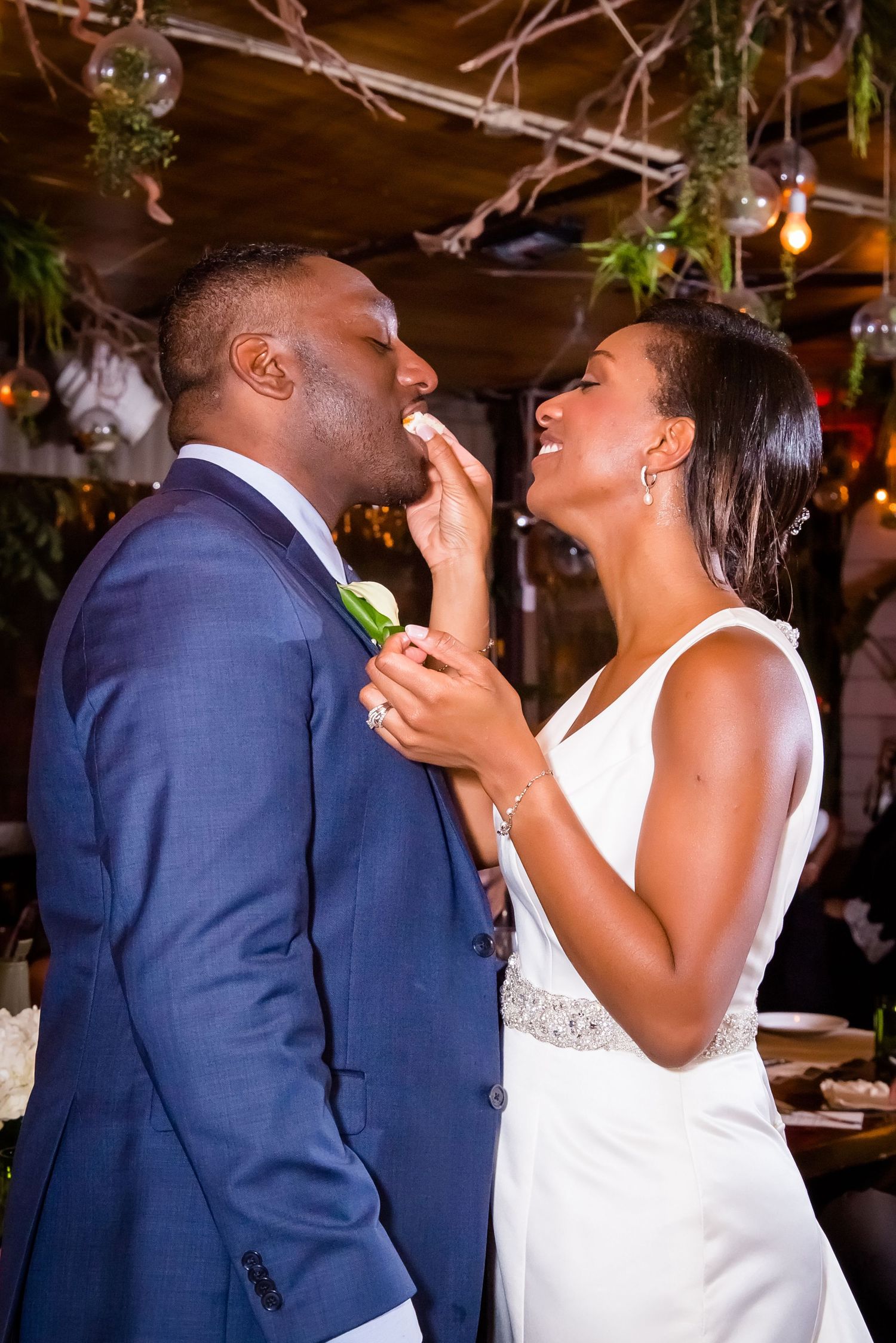 Couple laughing joyfully while feeding each other wedding cake during their reception celebration.