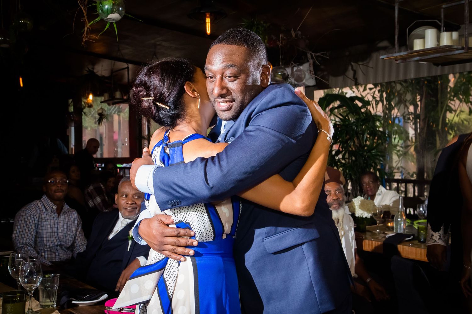Emotional embrace at a wedding reception in a dimly lit venue with festive decor.