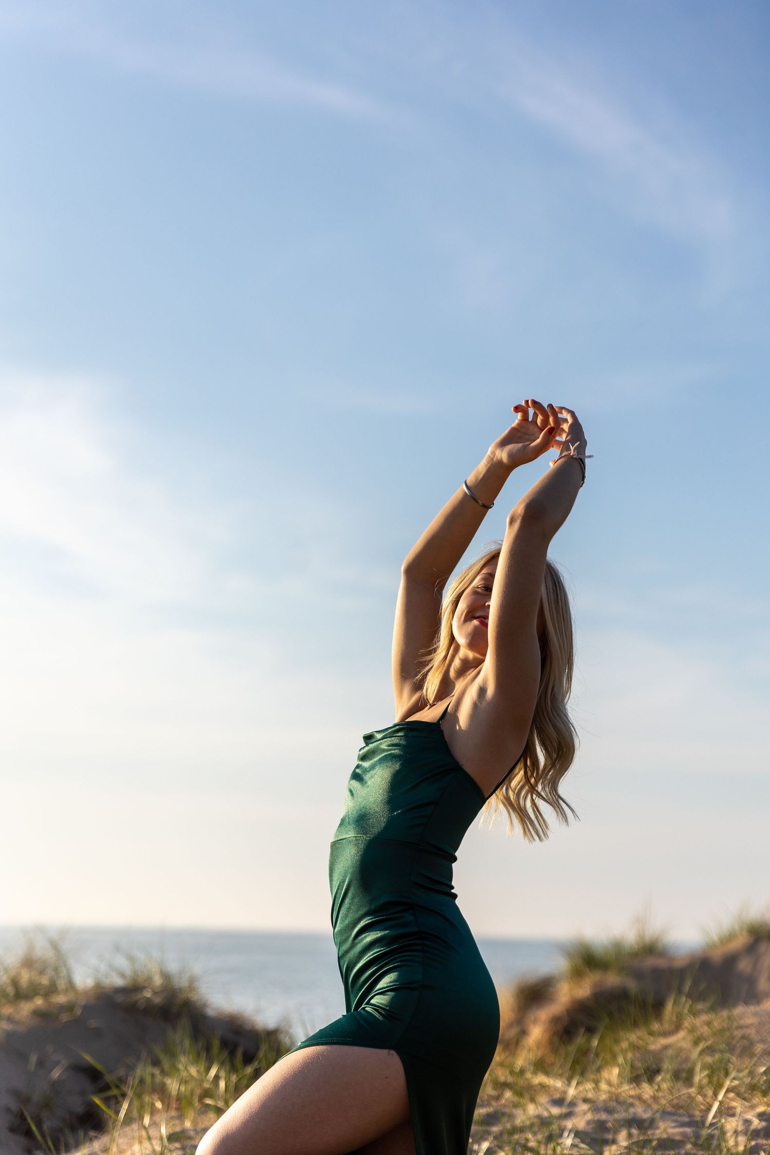 Blonde woman in elegant green dress posing on beach sand dunes with arms raised against blue sky.