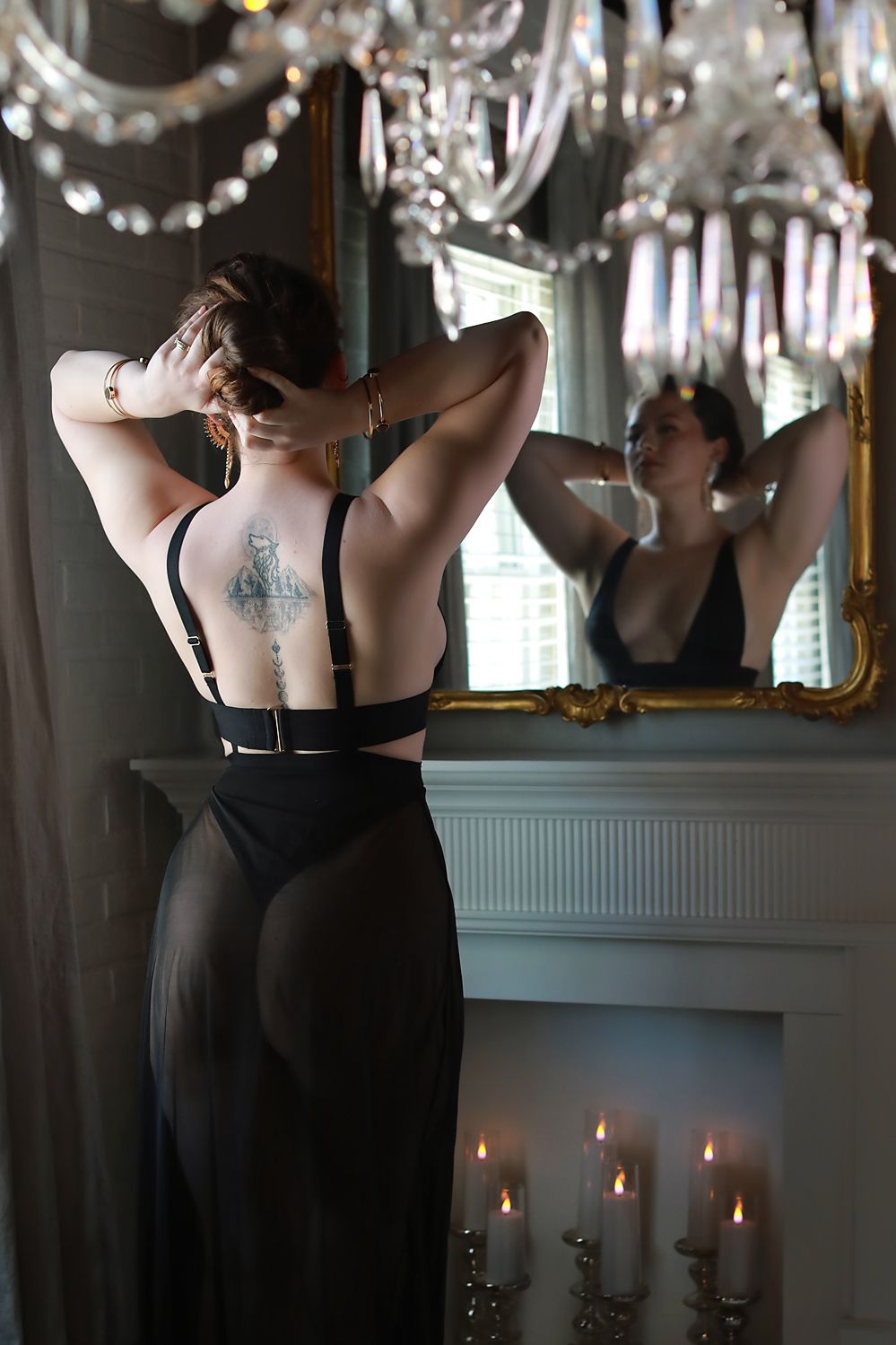 Figure in elegant black dress seen from behind by candlelit mirror with crystal chandelier overhead and reflective decor.