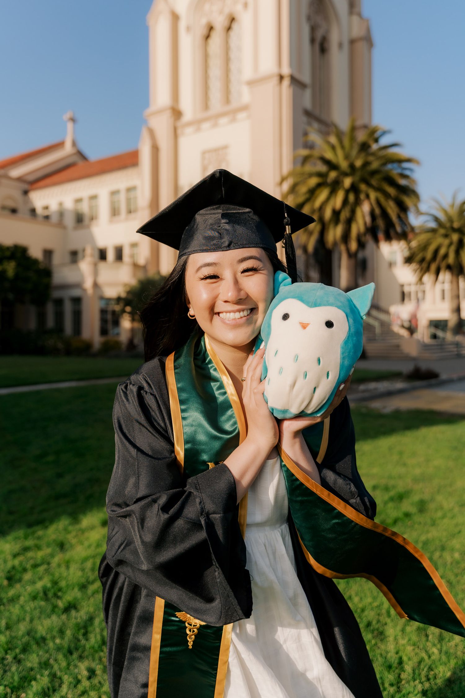 Graduate in cap and gown holds blue plush owl mascot in front of campus church.