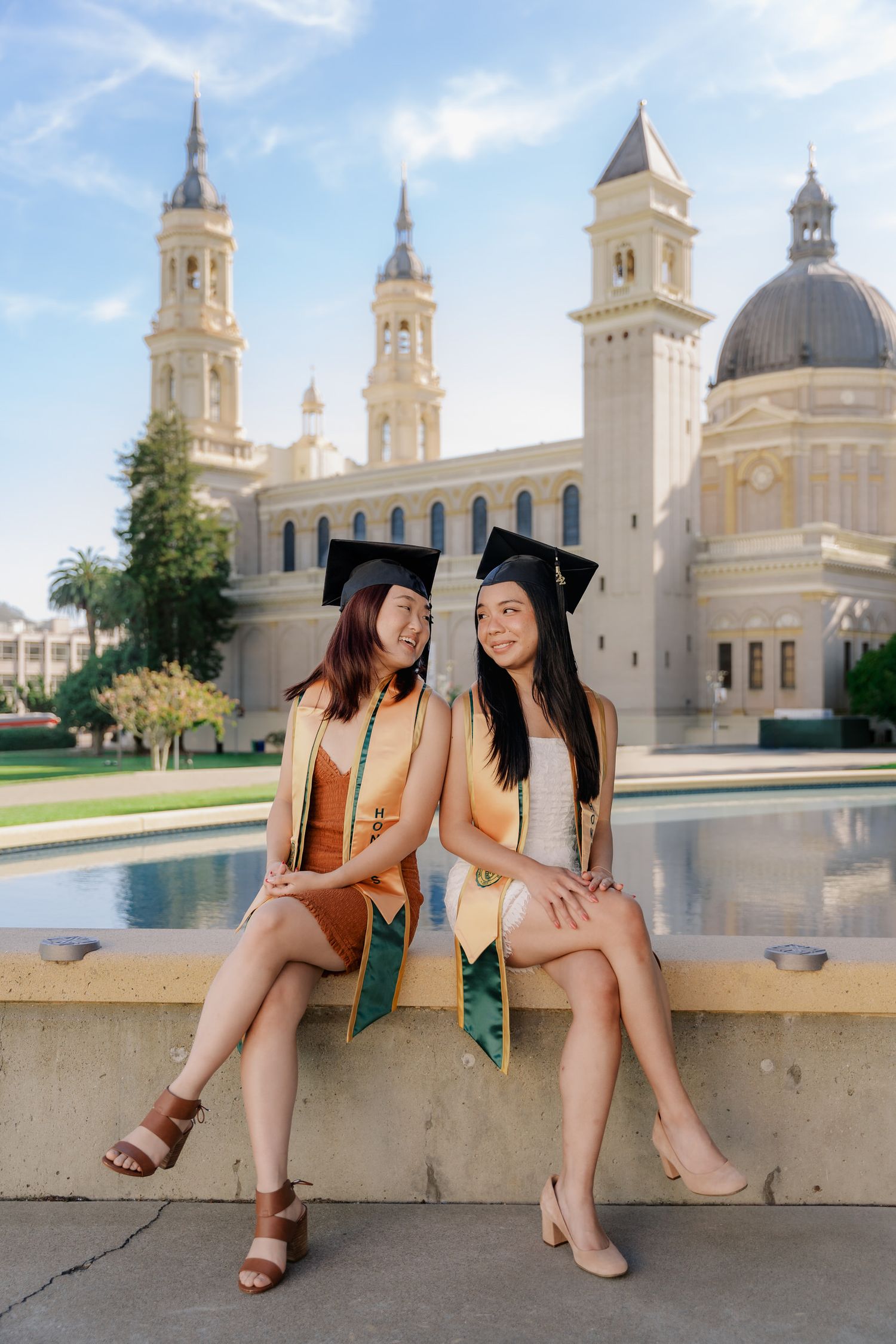 Two graduates in caps and gowns sit by fountain with ornate university building in background.