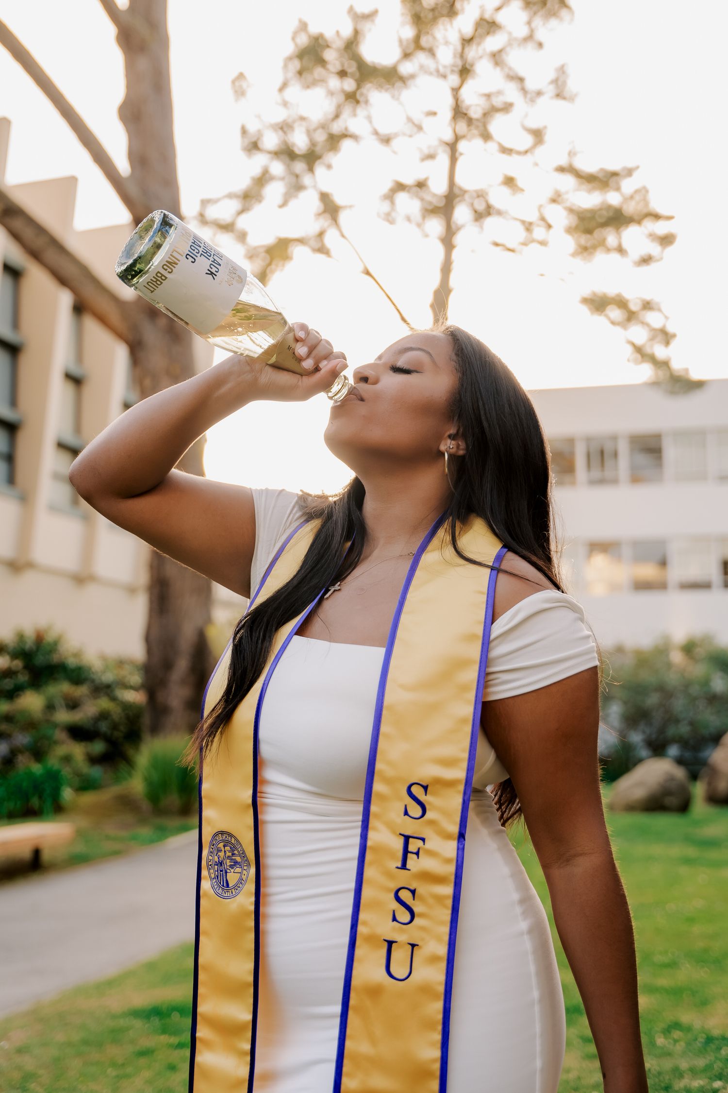 Graduate in white dress and graduation sash celebrates by drinking from champagne bottle.