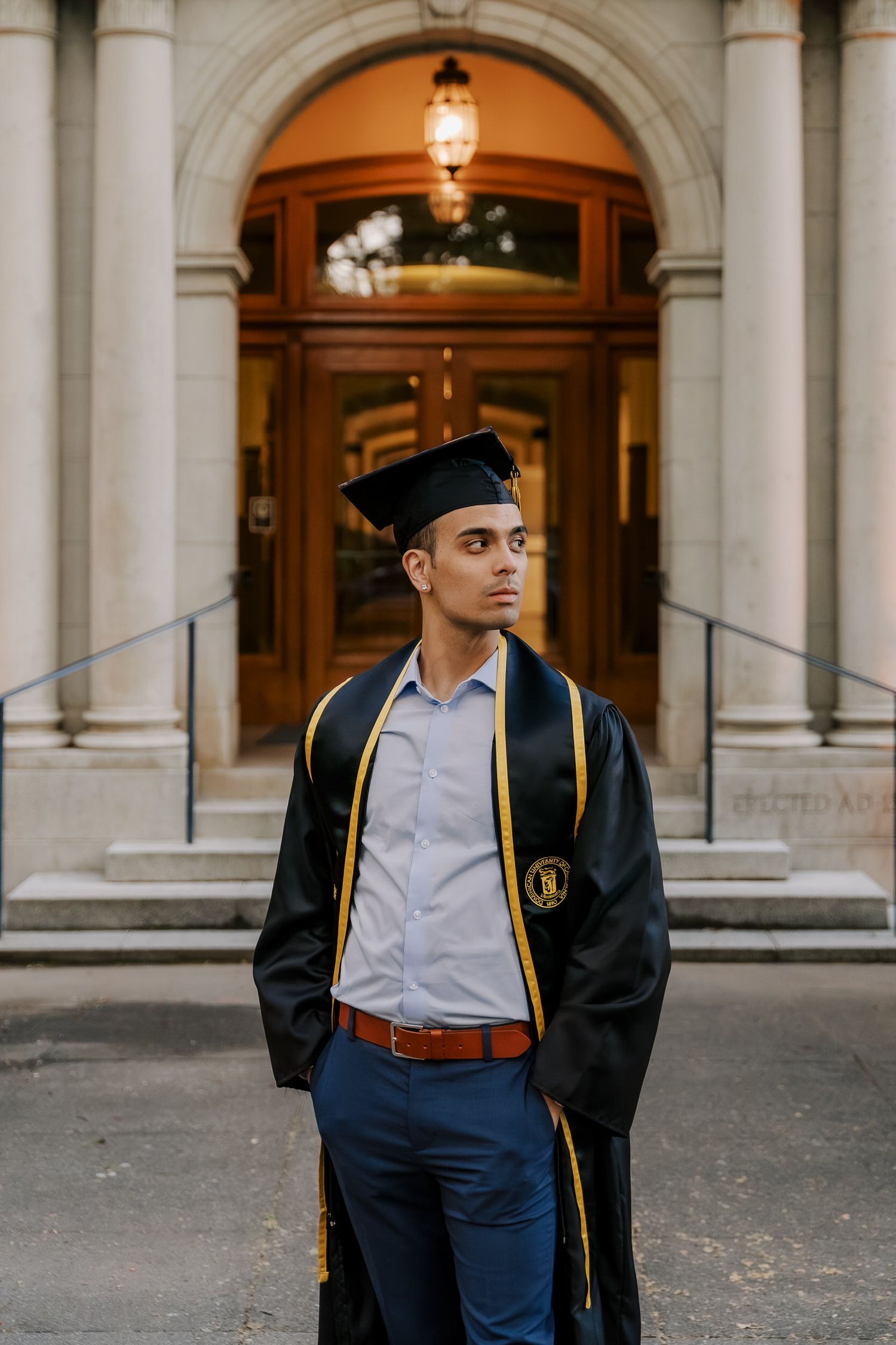 Graduate in formal attire and cap stands in front of ornate university entrance.