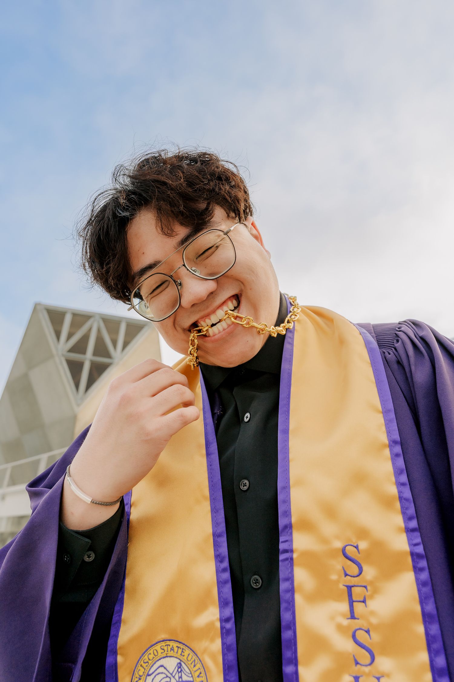 Graduate in academic regalia playfully bites graduation medal against modern architectural backdrop.