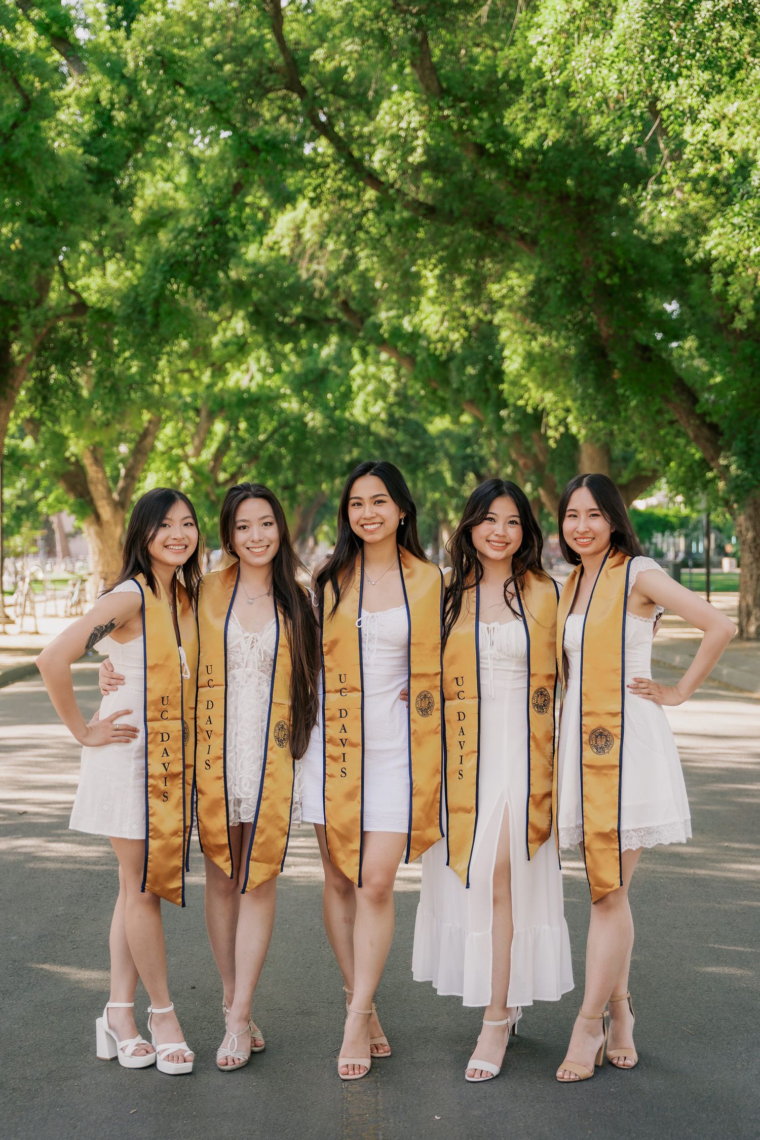 Graduate students in white dresses and gold stoles pose together on a tree-lined path during commencement.
