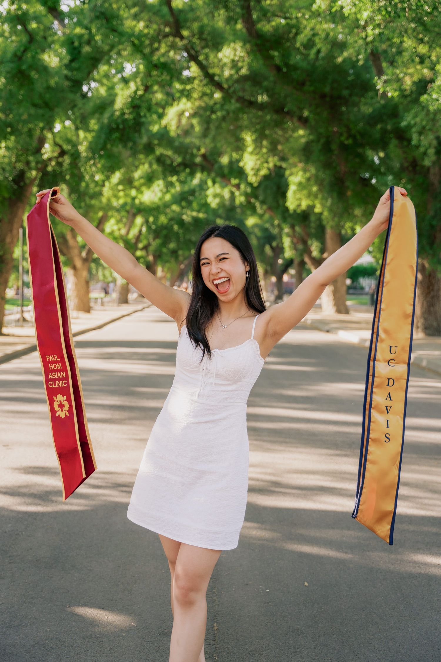 Graduate in white dress celebrates by holding up academic stoles on a sunny campus path.