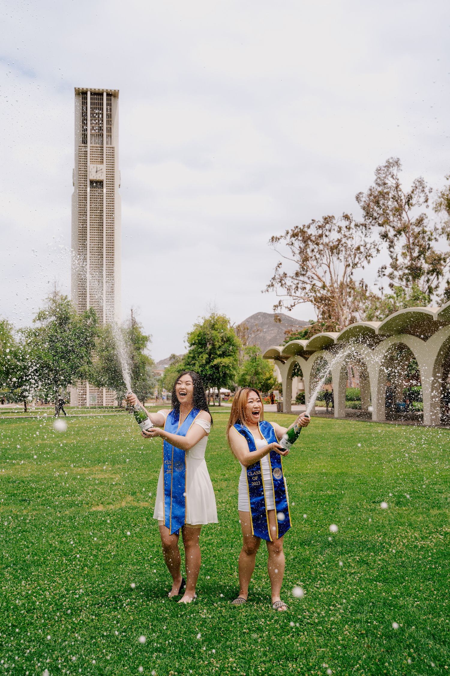 Two graduates spray champagne while celebrating on campus lawn with bell tower in background.