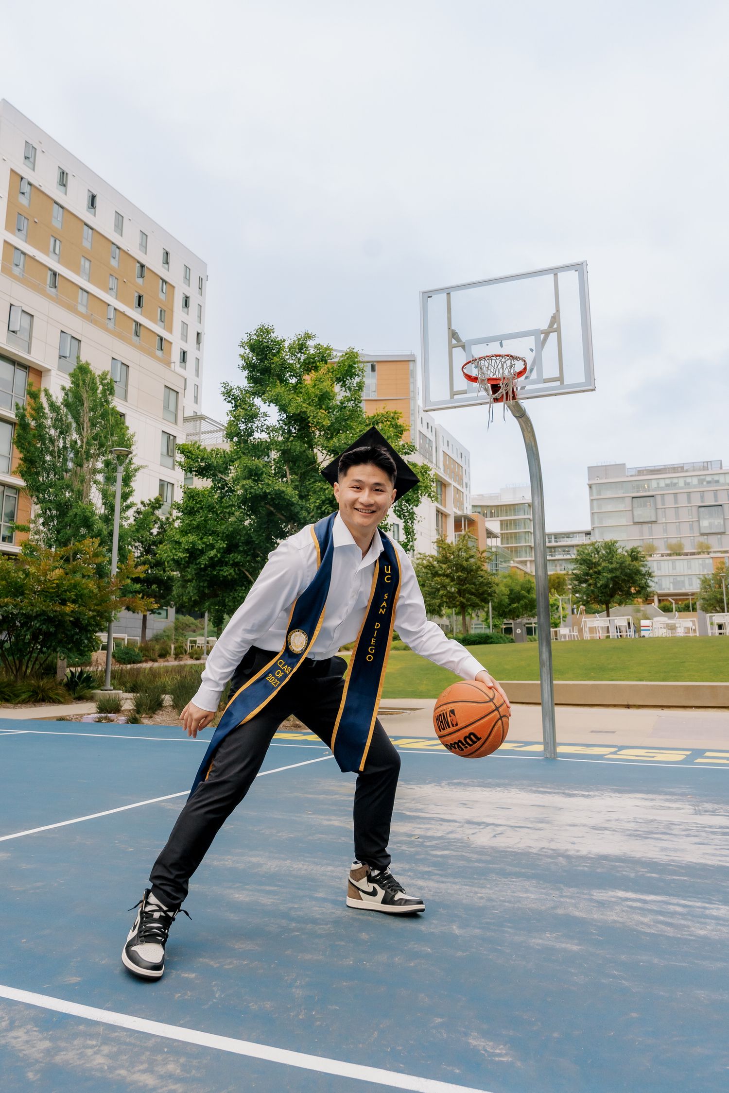 Graduate in academic regalia plays basketball on outdoor court.