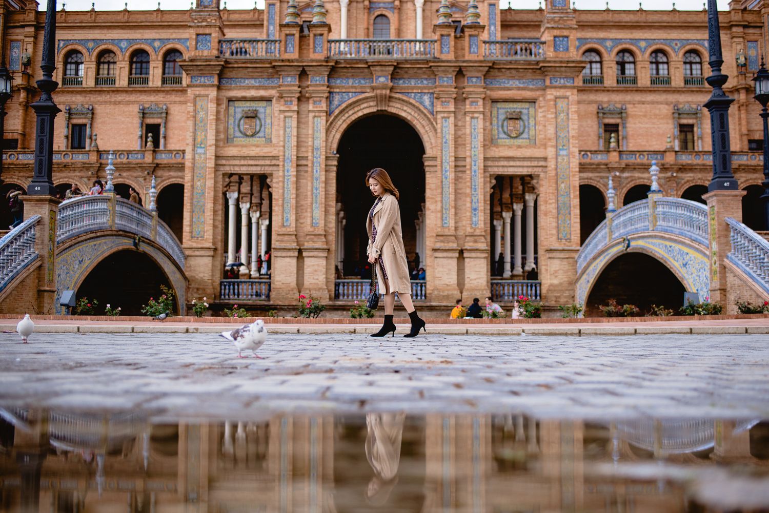 A person poses in front of ornate Spanish Renaissance architecture with water reflections.