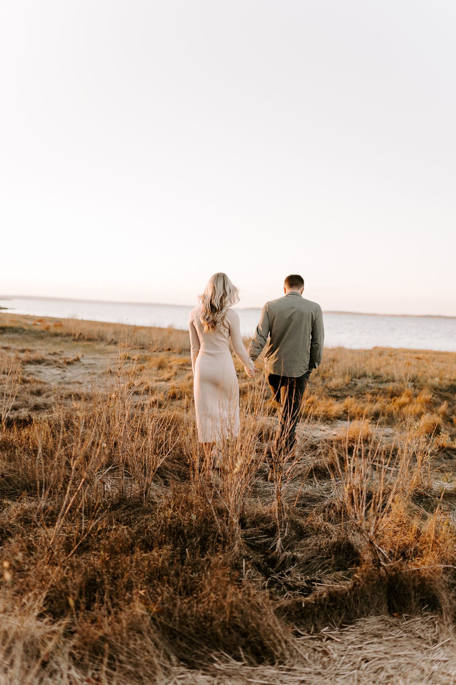 A couple walks hand in hand through a rustic field at dusk with warm sunlight behind them.