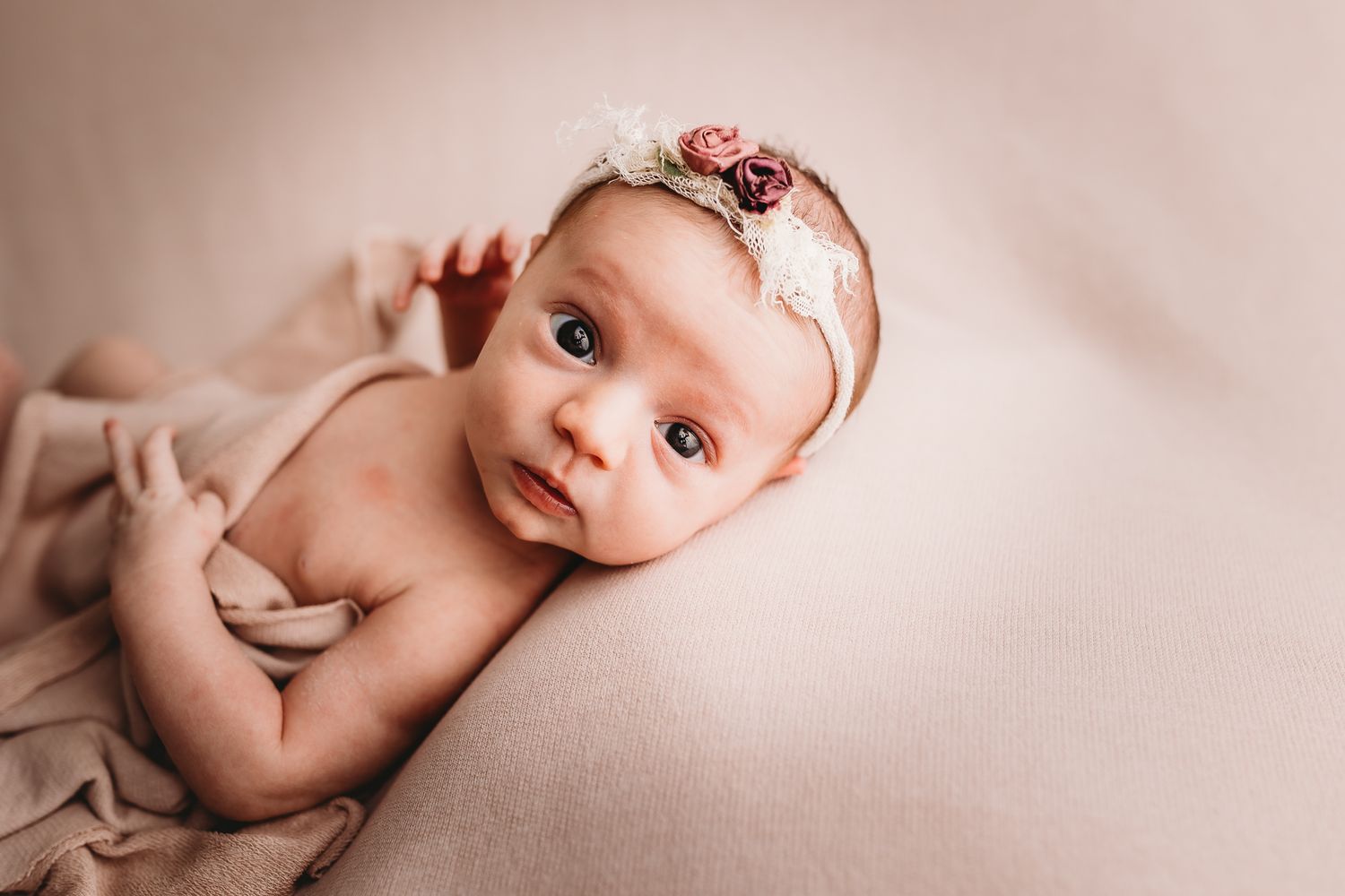 Adorable newborn baby girl wearing a floral headband lying on a soft beige blanket.