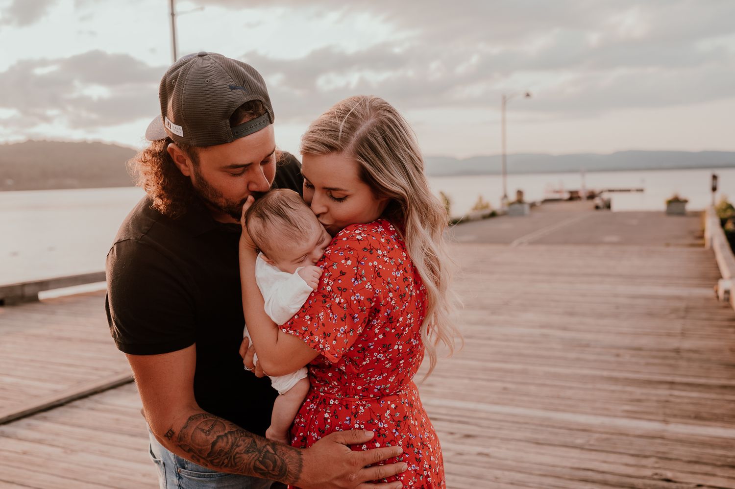 Tender moment shared between couple holding a baby on a wooden pier at sunset.