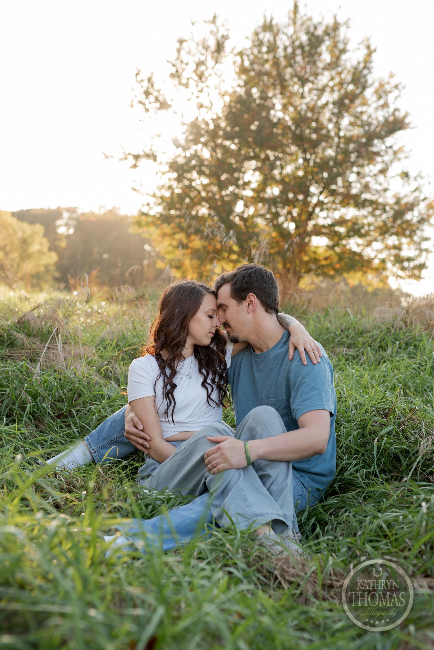 A romantic couple sits together in tall grass at sunset with a tree silhouetted in the background.
