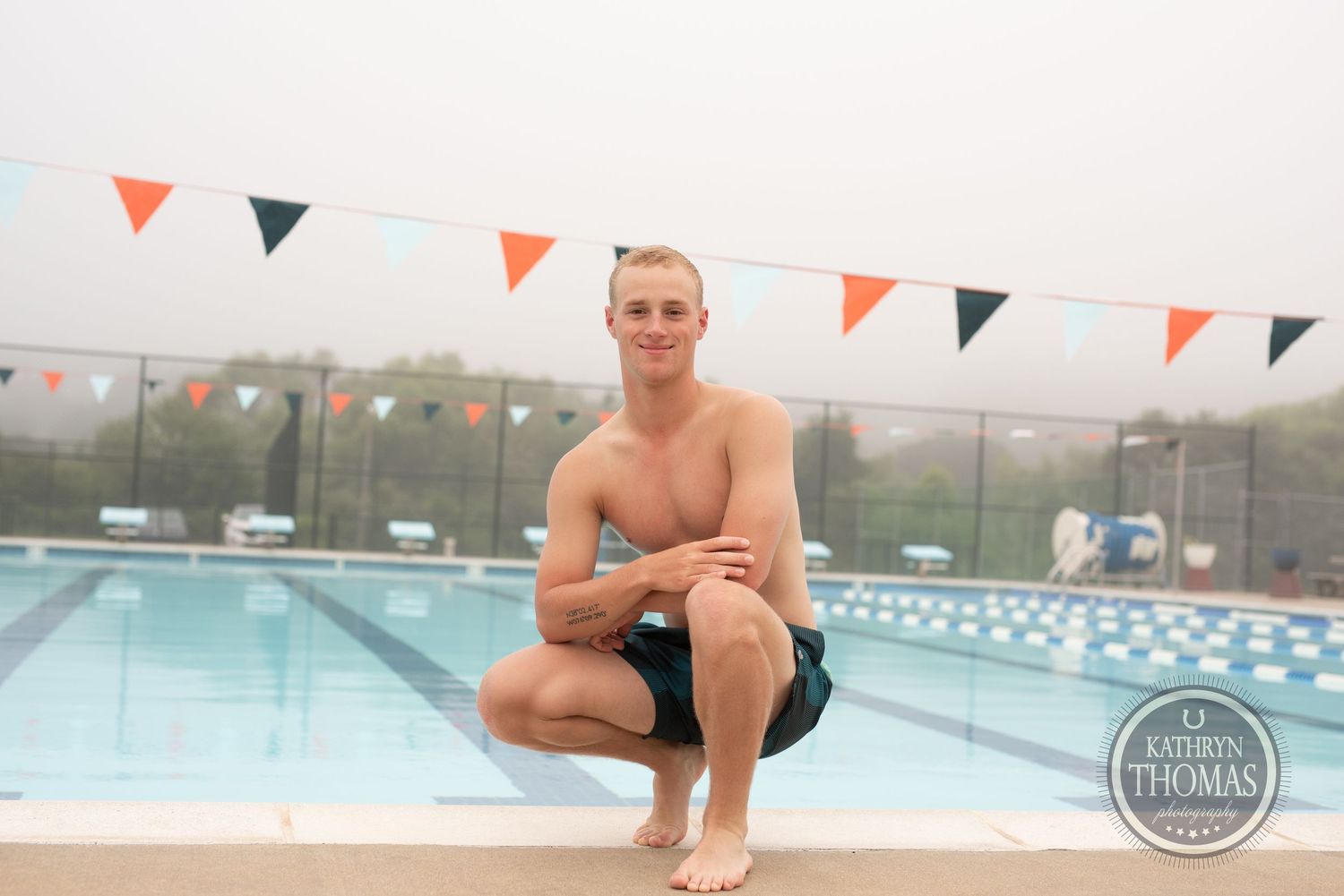 A swimmer crouches poolside at an outdoor swimming facility with colorful pennants decorating the area.