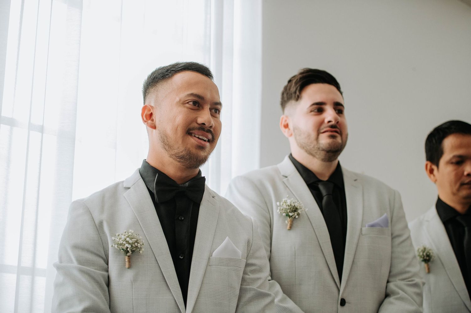 Three groomsmen in light colored suits smile during a wedding ceremony.