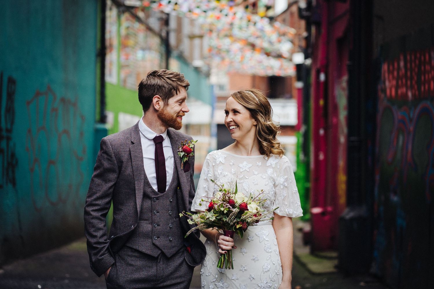 Newlyweds share an intimate moment in a colorful urban alley decorated with string lights.
