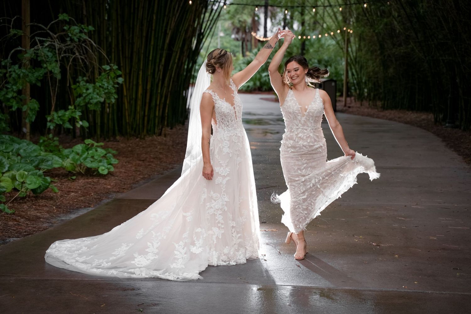 Two people in white wedding dresses dance joyfully on a wet pathway surrounded by greenery.