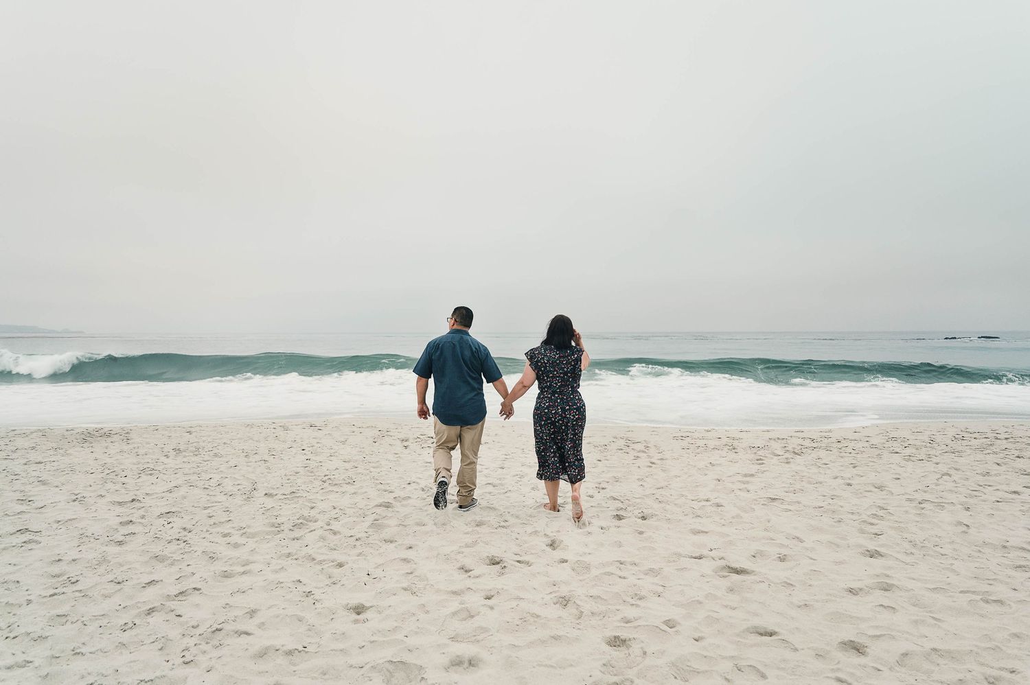 Couple walking hand in hand on a beach with waves crashing in the background.