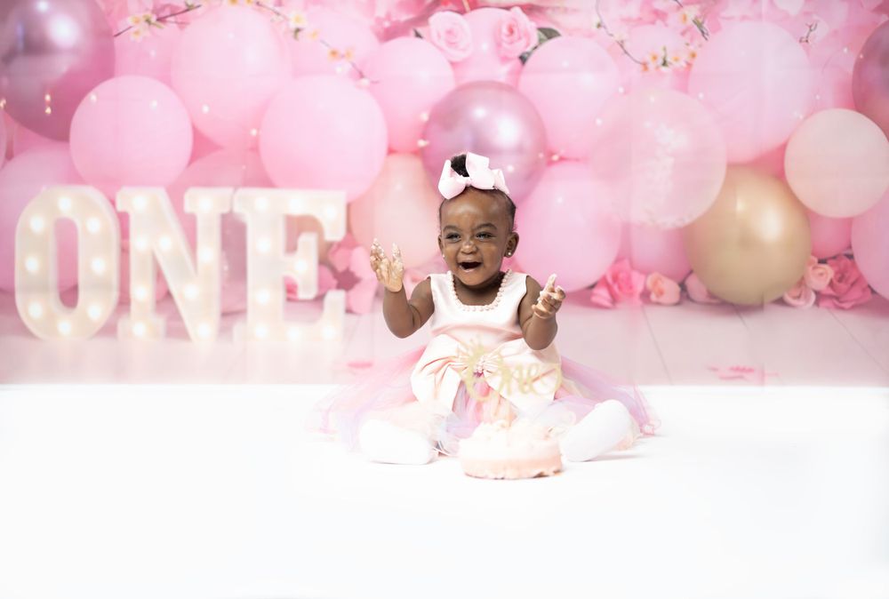 Smiling baby in tulle dress poses by ONE sign with pink balloon background.