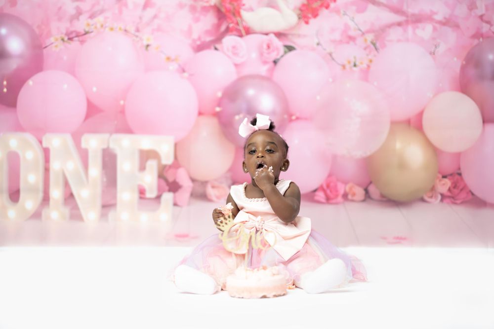 Baby poses playfully by ONE sign against pink balloon and floral backdrop.