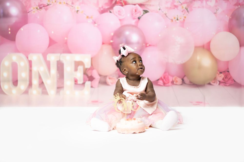 Baby in white dress sits beside illuminated ONE sign with festive pink balloon backdrop.
