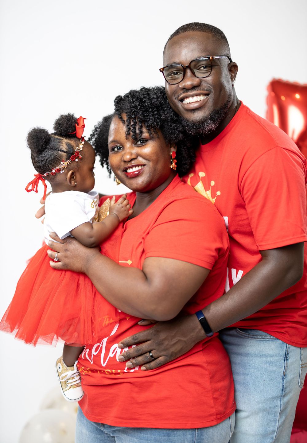 Family embraces in coordinated red outfits during professional photo session with white backdrop.