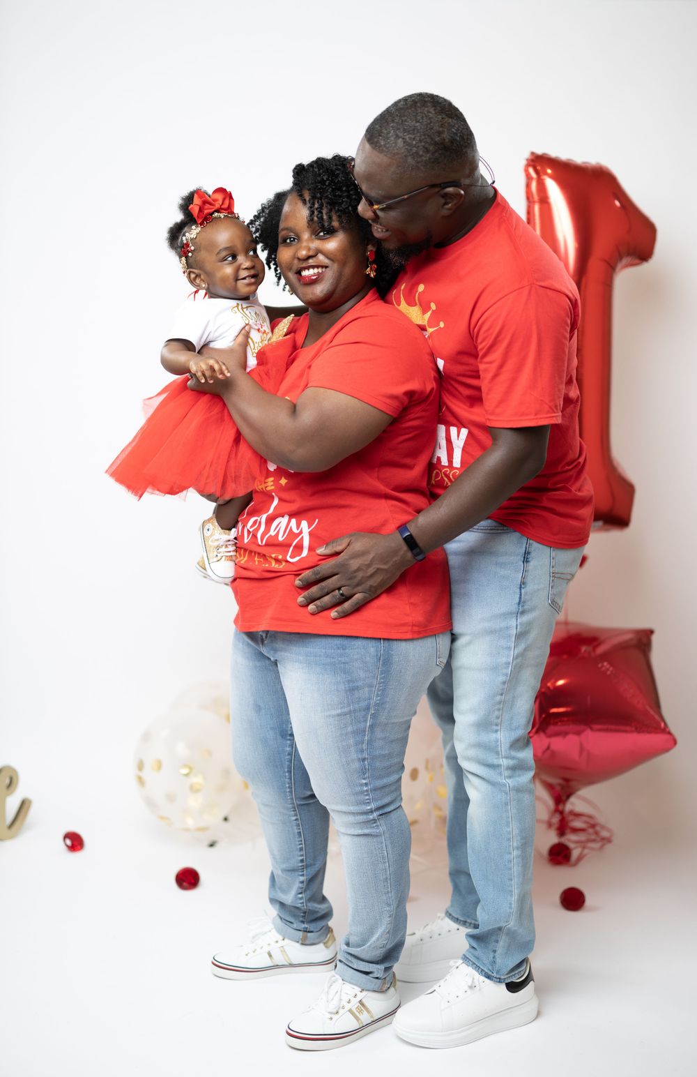 Family wearing red shirts poses together against white backdrop with balloons.