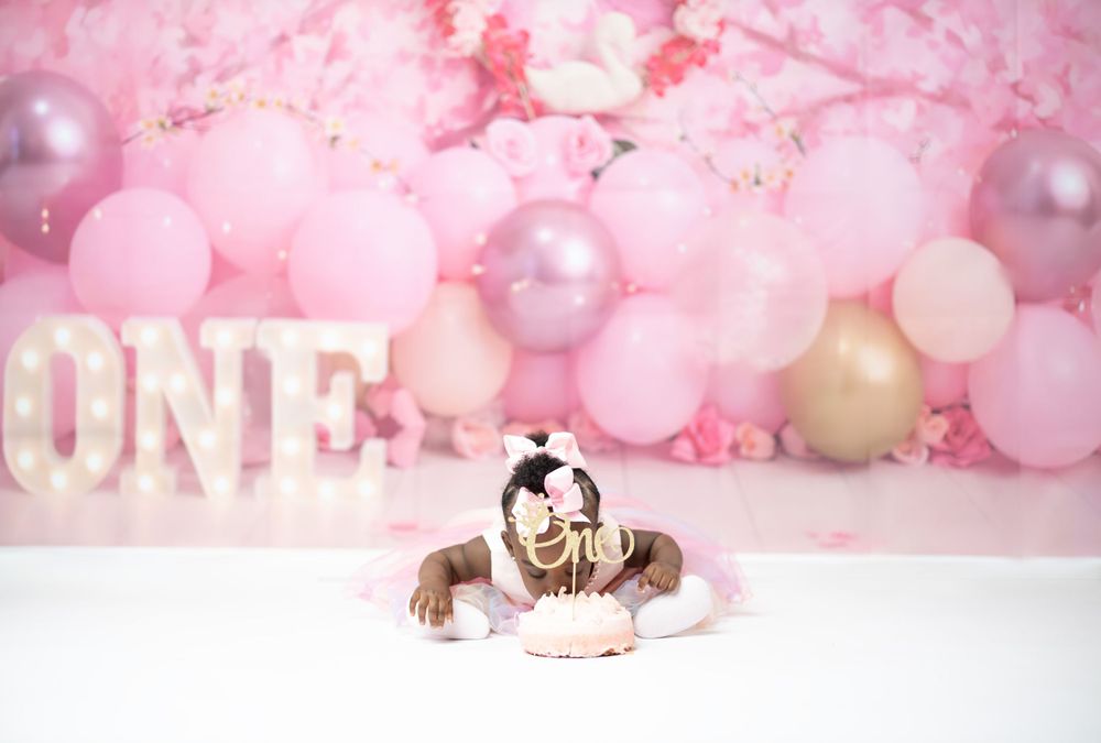Baby in white dress sits by illuminated ONE sign with pink balloon backdrop.