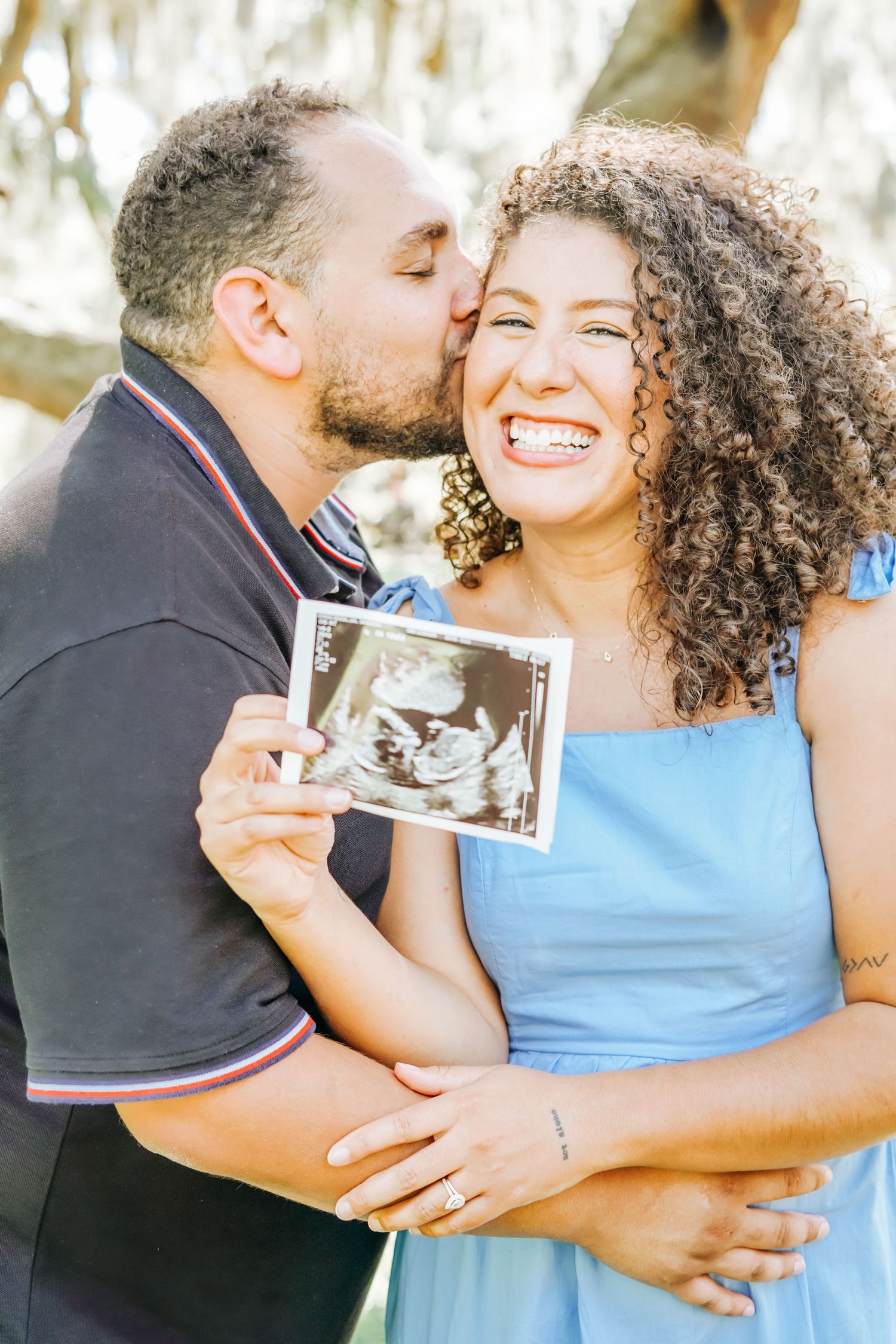 A couple sharing a joyful moment while holding an ultrasound photo in a park.