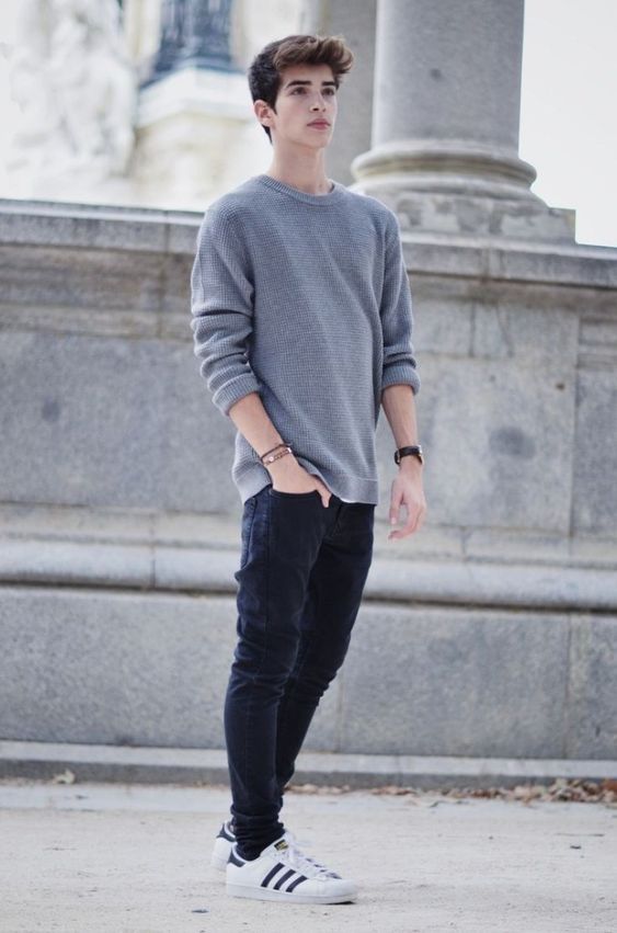 Casual street fashion featuring gray sweater, dark jeans and white Adidas sneakers by stone pillars.