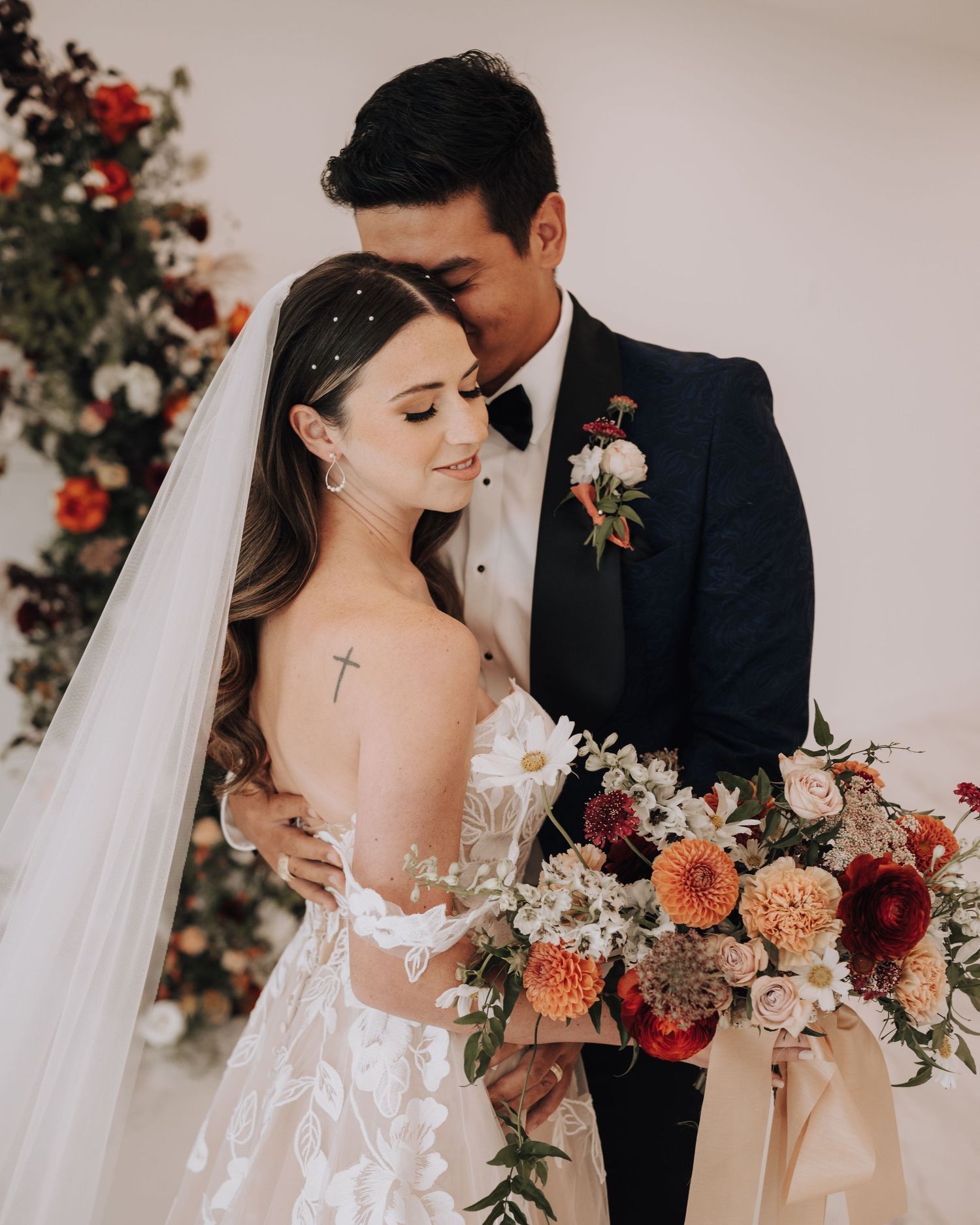 Intimate wedding portrait with couple and cross tattoo detail amid floral decorations.