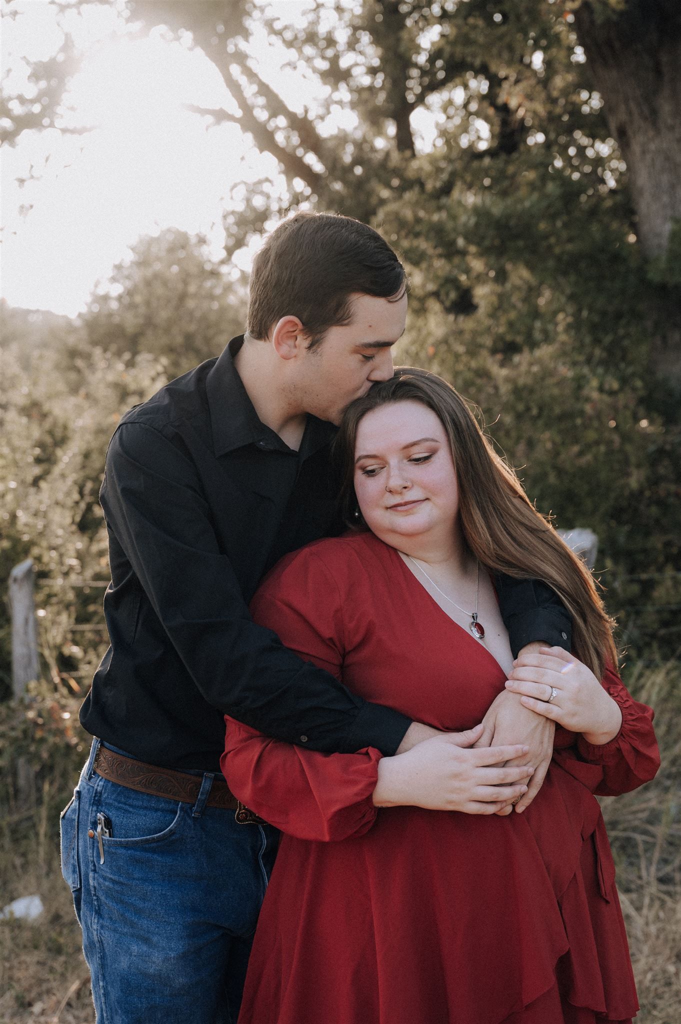 A couple shares a tender embrace in a natural setting with sunlight filtering through trees.