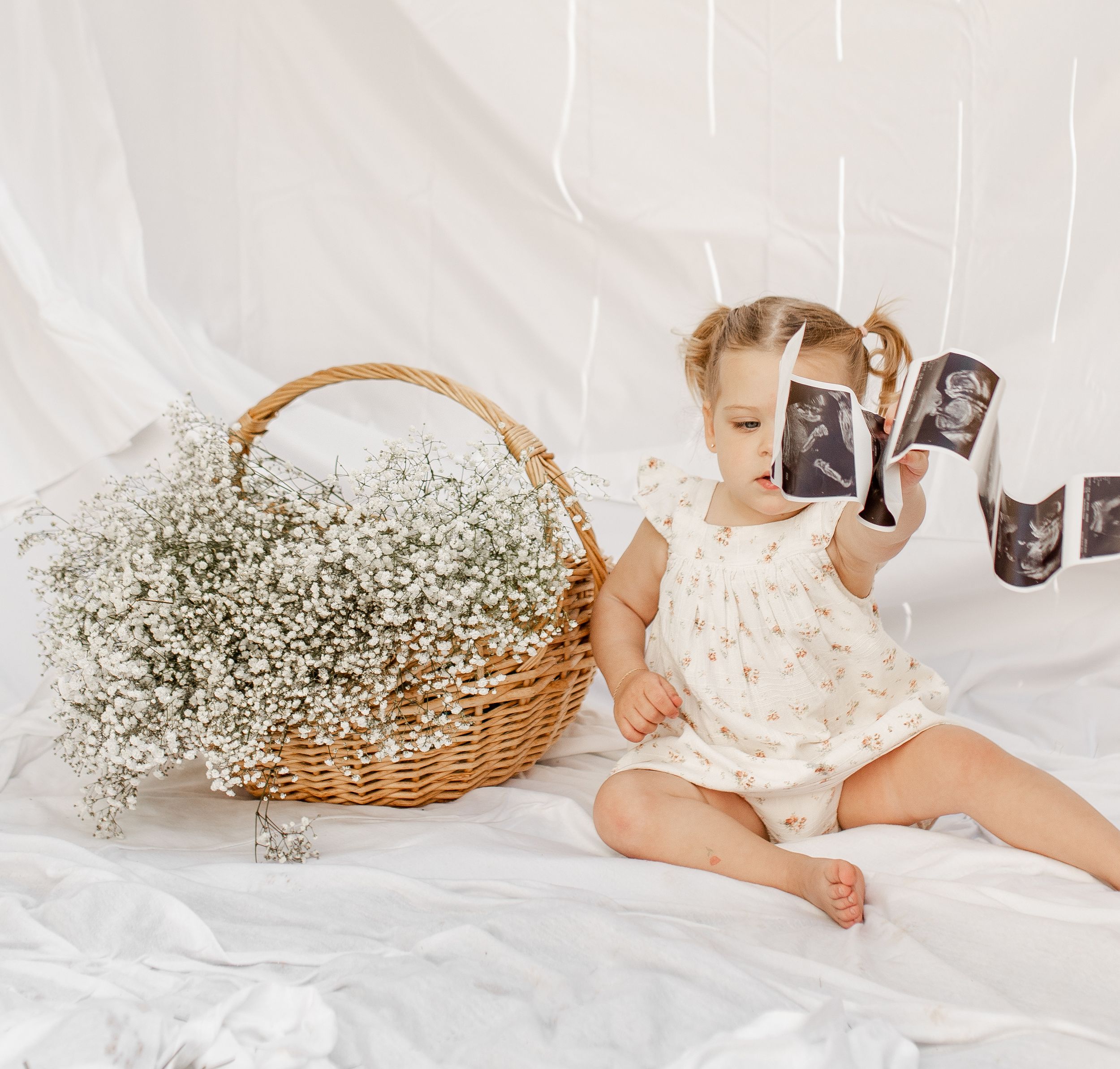 A toddler in white dress sits beside basket of baby's breath flowers examining ultrasound strips.