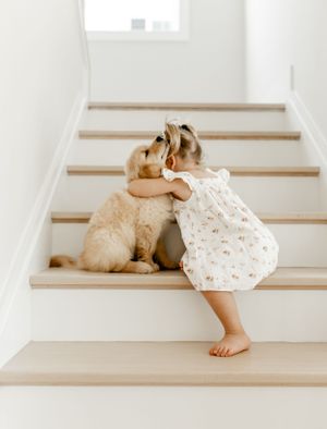 A young child in a floral dress hugging a golden retriever puppy on wooden stairs.