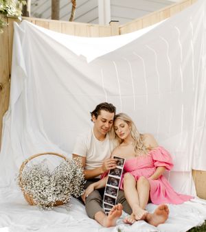 Couple sitting on white blanket outdoors holding ultrasound photos near baby's breath flowers.