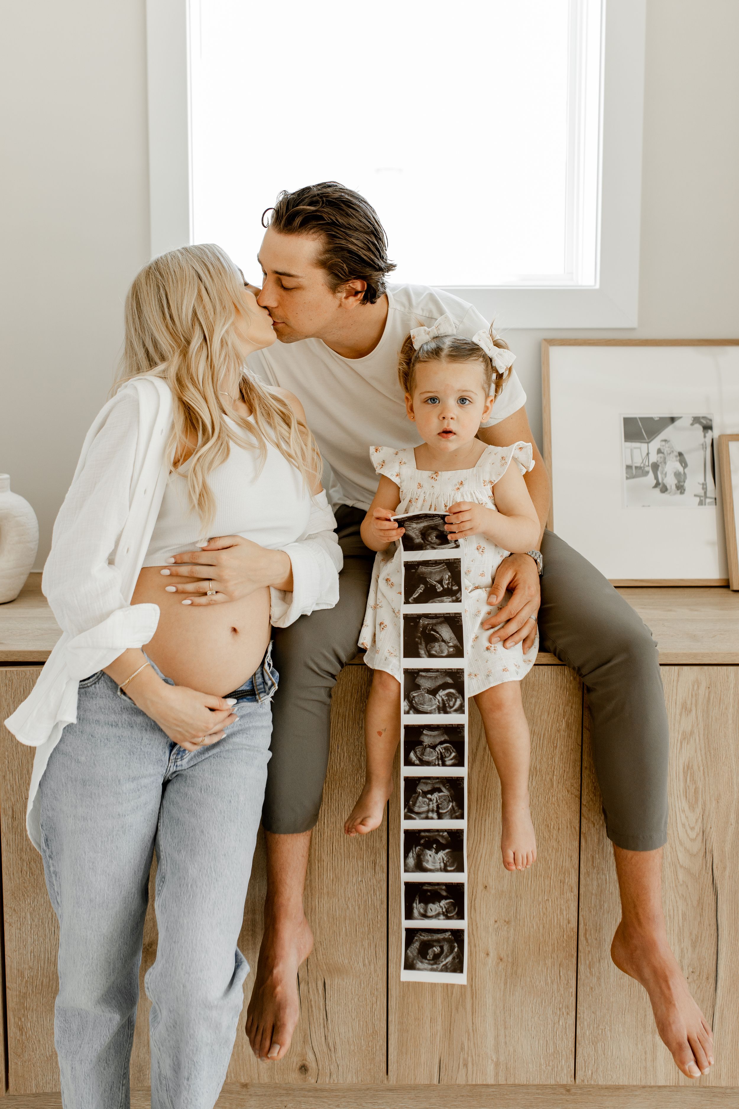 A sweet pregnancy announcement photo featuring ultrasound images and a toddler sitting on wooden cabinets.
