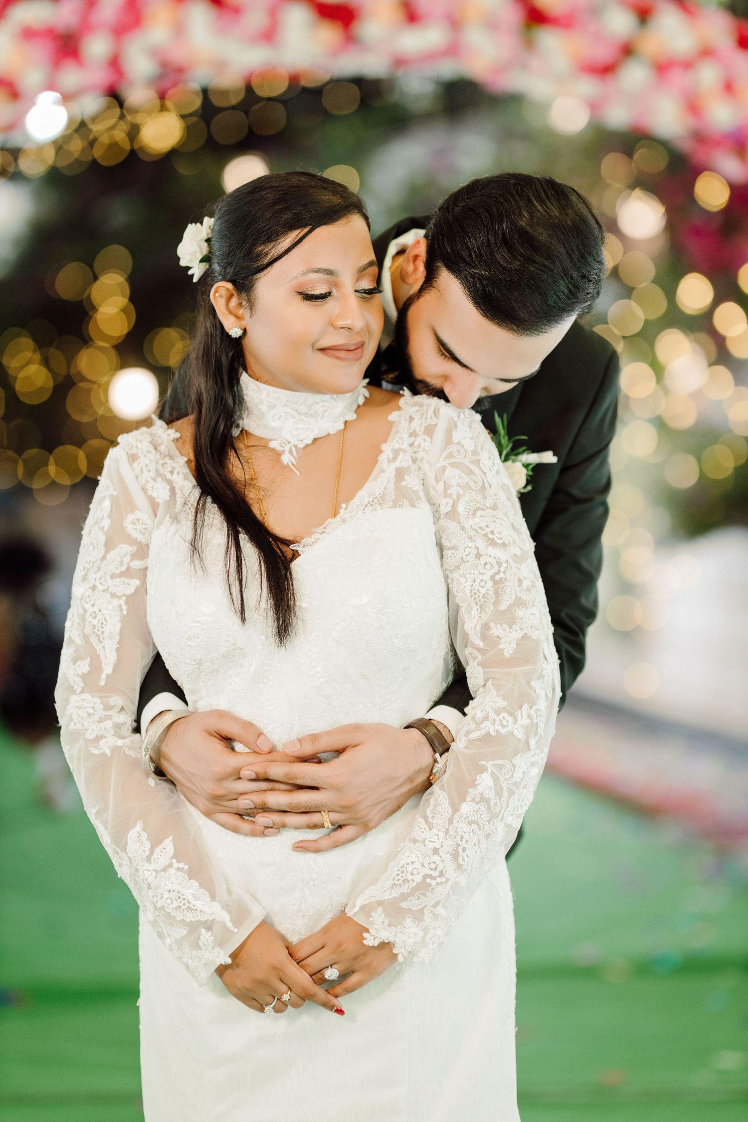 A romantic wedding couple embraces in a garden setting with bokeh lights and lace dress details.