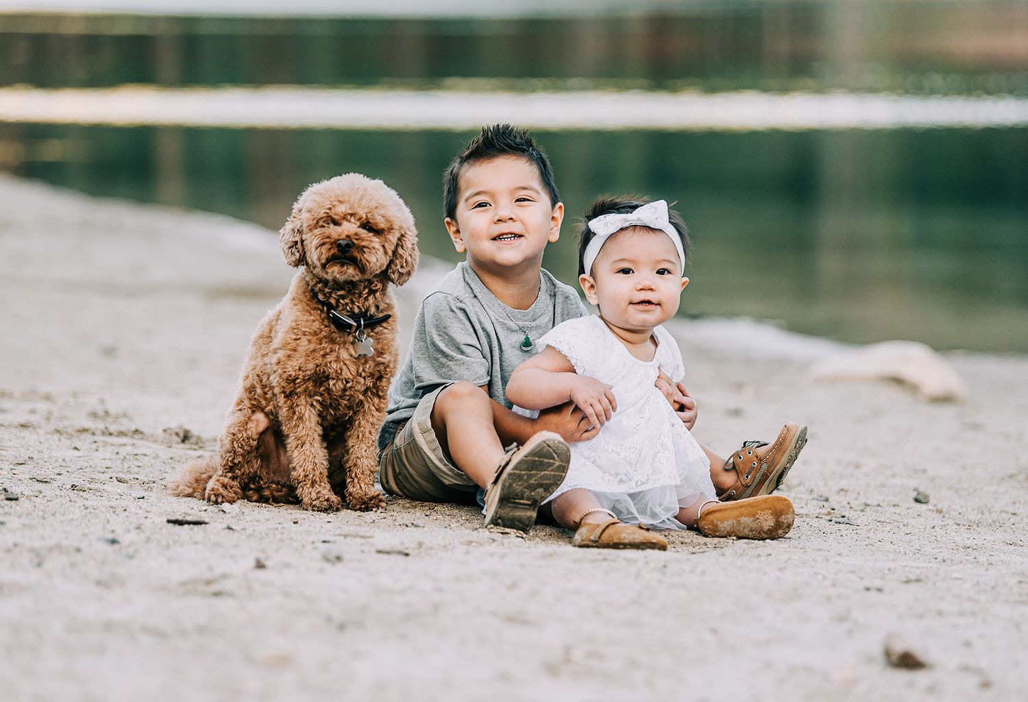 Children sit with their fluffy brown poodle by a serene lake.