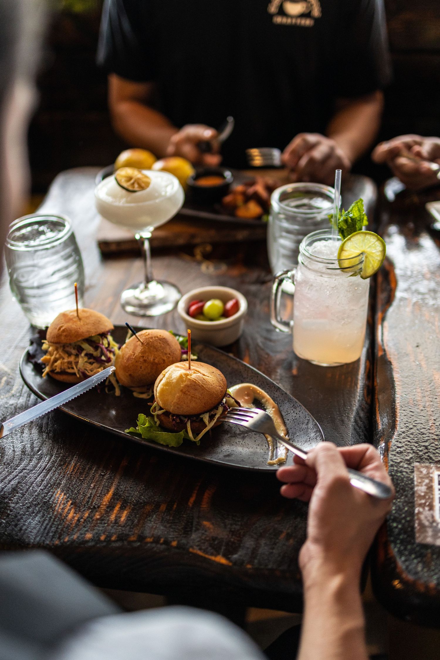 Gourmet sliders served with cocktails and garnishes on rustic wooden table.