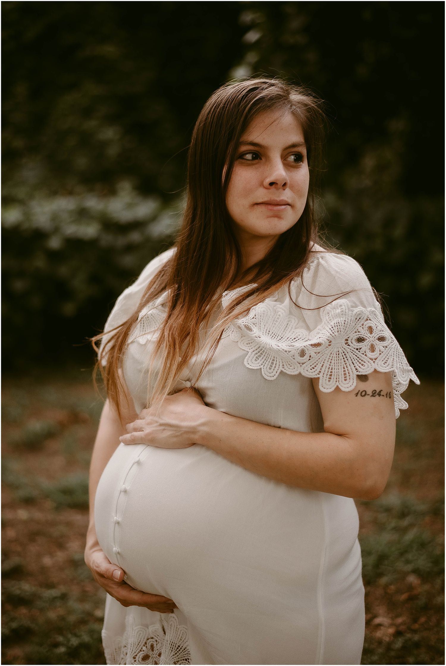 Close-up maternity portrait of pregnant woman wearing white lace dress in outdoor garden setting.