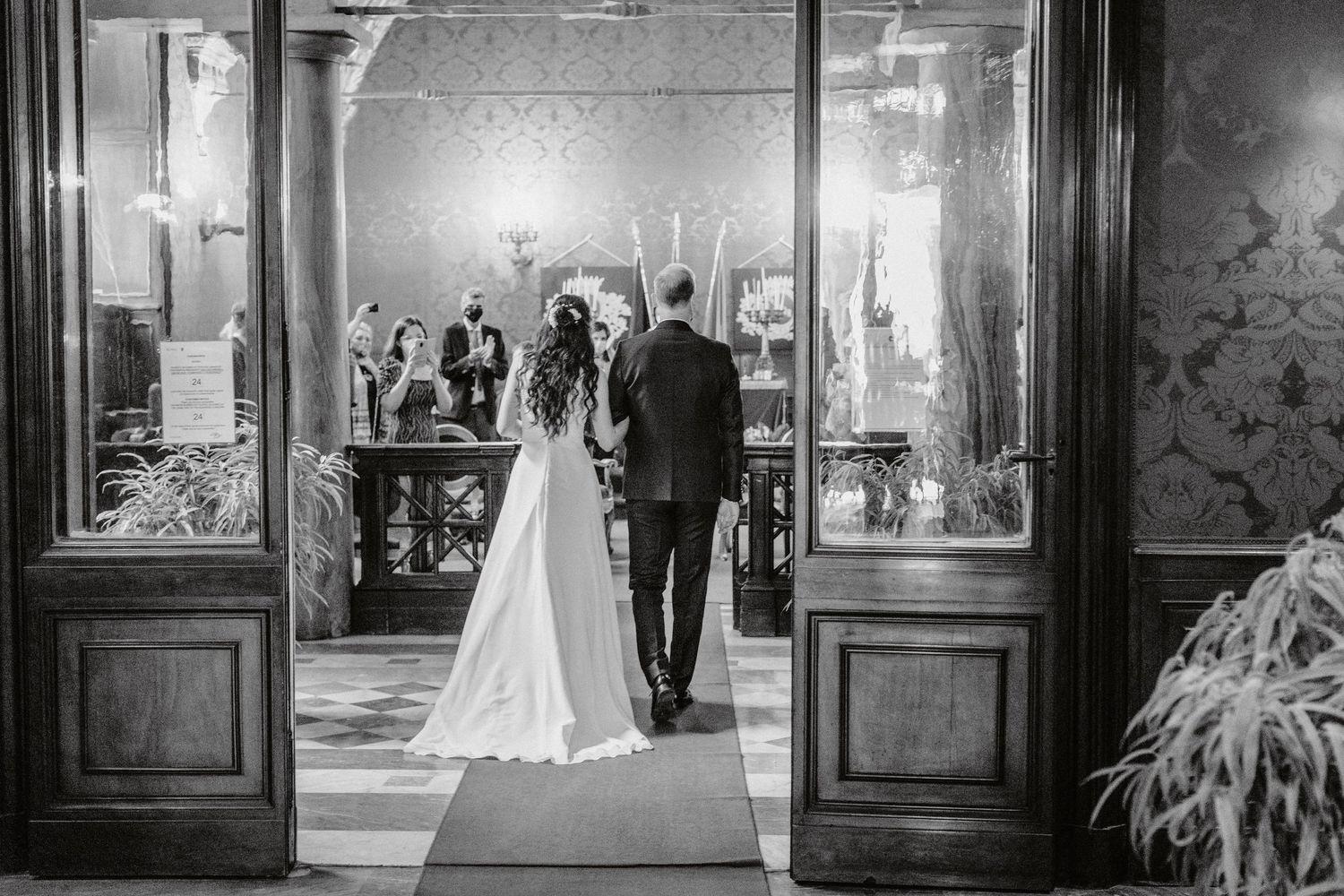 Black and white wedding photography sequence showing couple walking through ornate venue hallway.