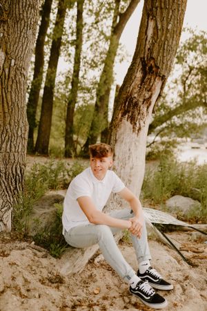 Person relaxes on sandy ground near trees wearing white shirt and jeans with black sneakers.