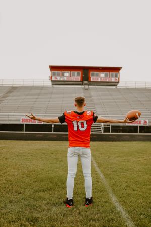 Football player in orange jersey number 10 stands with arms outstretched on empty field with stadium bleachers.
