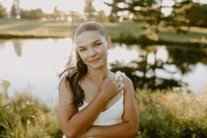 Person in white dress poses outdoors near pond during golden hour.