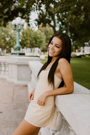 Person in beige dress leaning against stone railing in park setting.