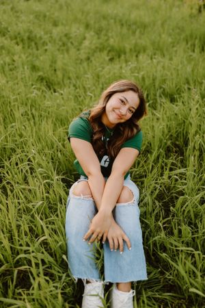 Young person in green shirt sitting casually in tall grass field during sunset.
